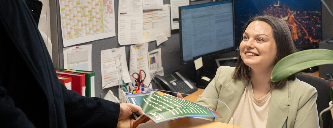 Die Sekretariatsmitarbeiterin sitzt am Schreibtisch und nimmt eine Mappe von einem Schüler entgegen.