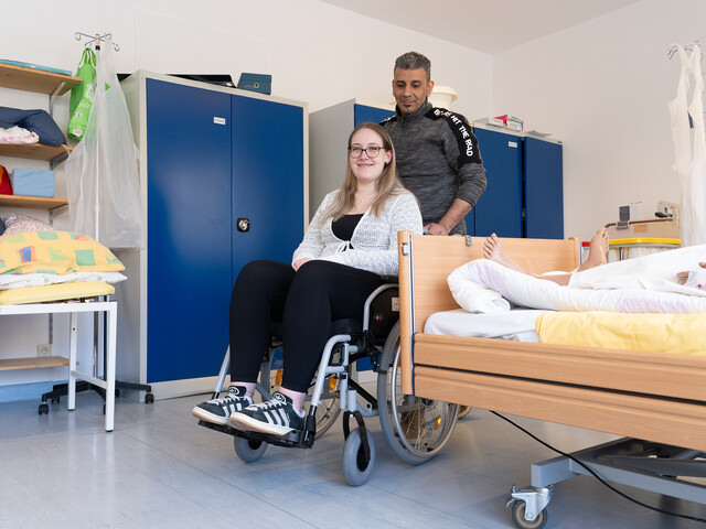 Eine Schülerin sitzt im Rollstuhl, ein Schüler schiebt sie neben dem Krankenbett vorbei.