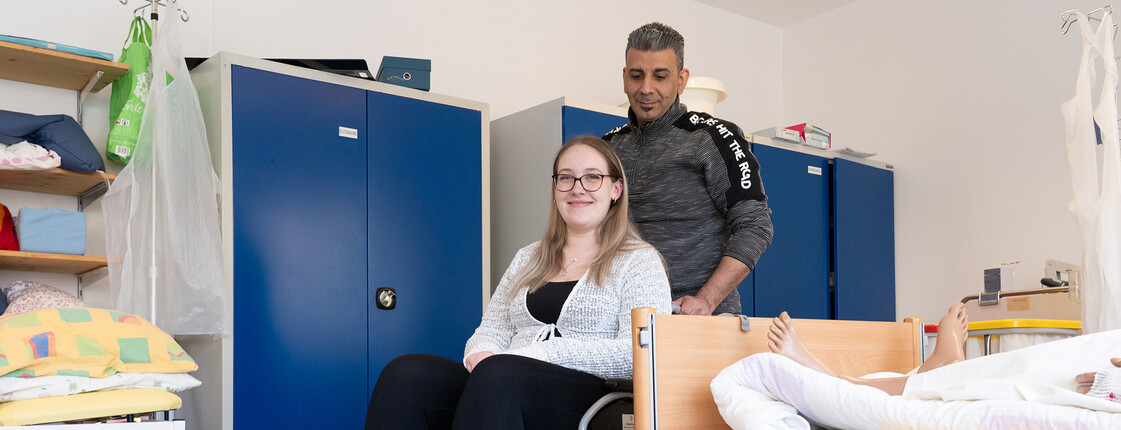 Eine Schülerin sitzt im Rollstuhl, ein Schüler schiebt sie neben dem Krankenbett vorbei.