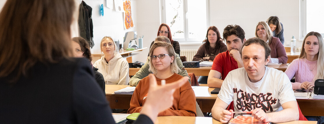 Eine Lehrerin steht vor der Klasse und unterrichtet, die unterschiedlich alten Studierenden sitzen auf den Bänken.