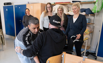 Ein Schüler setzt einen anderen Schüler auf einen Stuhl. Seine Mitschüler*innen beobachten dies.