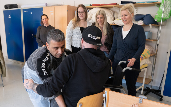 Ein Schüler setzt einen anderen Schüler auf einen Stuhl. Seine Mitschüler*innen beobachten dies.