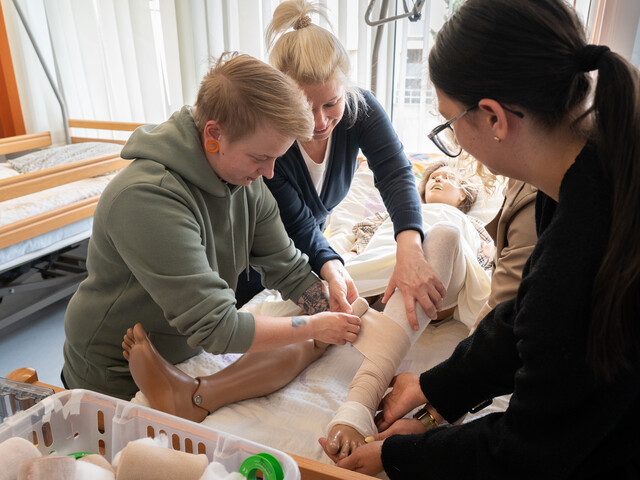 Eine Schülerin bringt einen Verband auf einer Puppe an, eine Lehrkraft hilft ihr dabei.