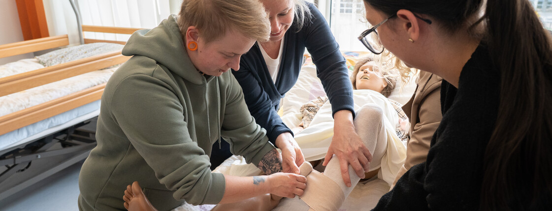 Eine Schülerin bringt einen Verband auf einer Puppe an, eine Lehrkraft hilft ihr dabei.