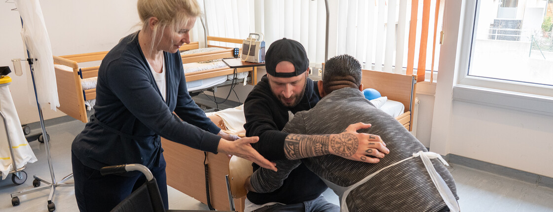 Ein Schüler hebt einen anderen Schüler von einem Rollstuhl auf einen Stuhl. Eine Lehrerin assistiert ihm dabei.