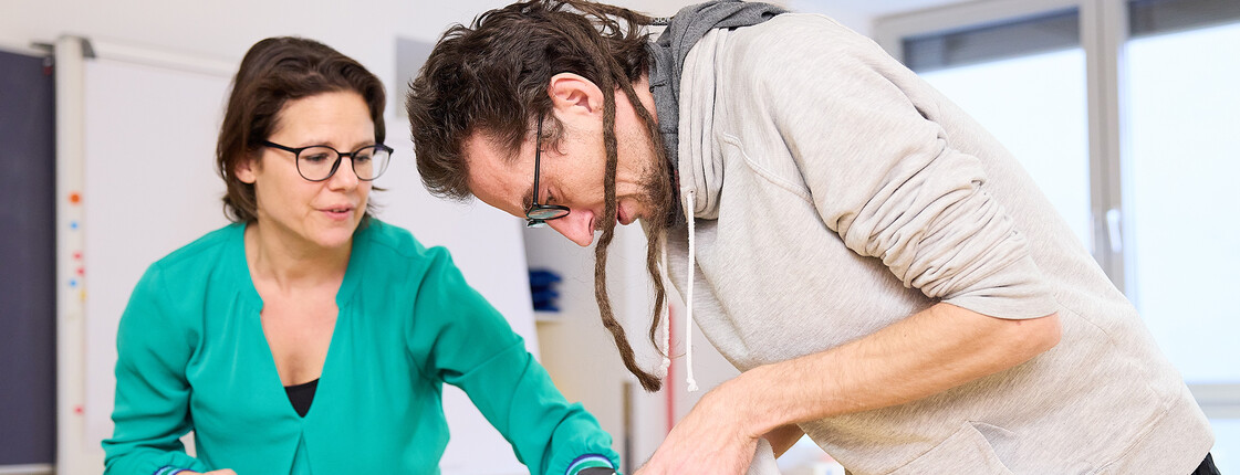 Schüler übt das fachgerechte Anlegen eines Fußverbands im Schulungsraum, unter Aufsicht einer Lehrkraft.