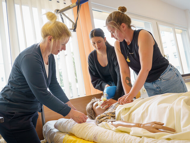 Zwei Schüler*innen üben unter Aufsicht der Lehrkraft Herzdruckmassage und Beatmung an einer Übungspuppe im Pflegebett.