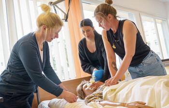 Zwei Schüler*innen üben unter Aufsicht der Lehrkraft Herzdruckmassage und Beatmung an einer Übungspuppe im Pflegebett.
