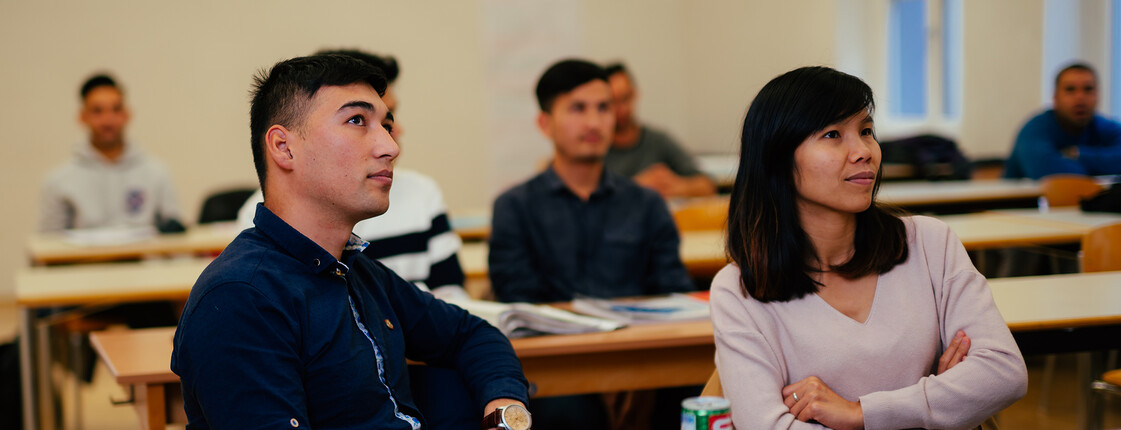 Studierende sitzen auf Schulbänken und sehen auf die Tafel