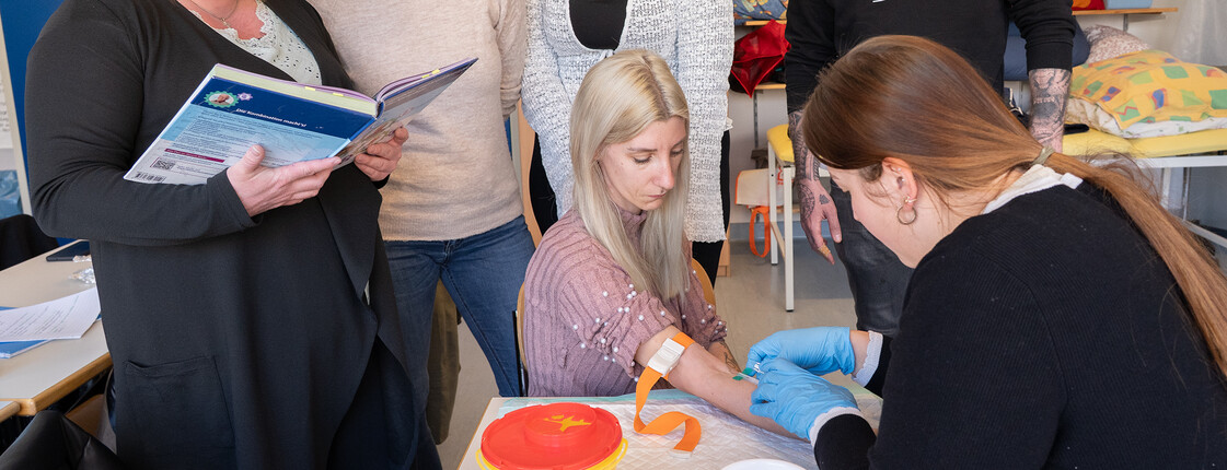 Eine Schülerin nimmt bei einer Mitschülerin Blut ab, die Utensilien stehen auf einem Tisch. Mitschüler*innen sehen ihr dabei zu.