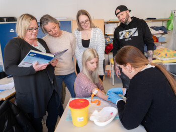 Eine Schülerin nimmt bei einer Mitschülerin Blut ab, die Utensilien stehen auf einem Tisch. Mitschüler*innen sehen ihr dabei zu.