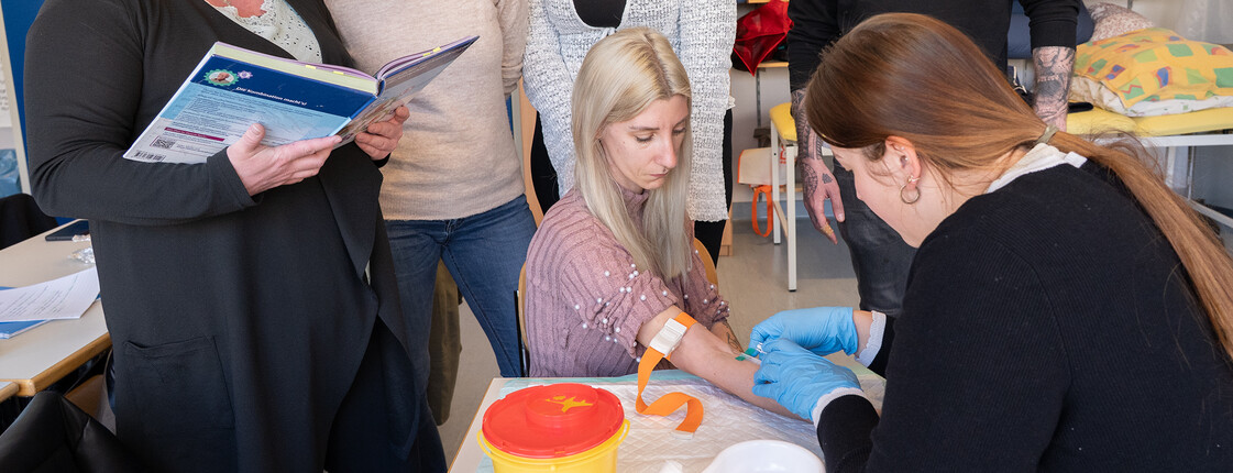 Eine Schülerin nimmt bei einer Mitschülerin Blut ab, die Utensilien stehen auf einem Tisch. Mitschüler*innen sehen ihr dabei zu.