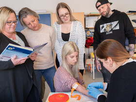 Eine Schülerin nimmt bei einer Mitschülerin Blut ab, die Utensilien stehen auf einem Tisch. Mitschüler*innen sehen ihr dabei zu.
