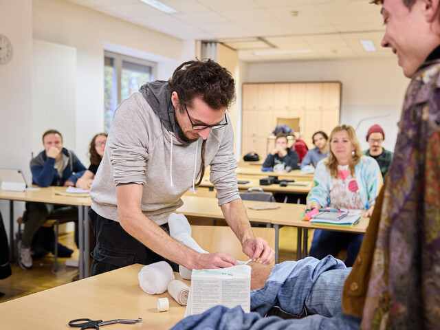Schüler wickelt Verband um Fuß, während andere im Klassenraum zuschauen.