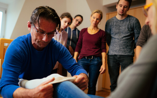 Ein Studierender zieht einer Mitschülerin einen Kompressionsstrumpf an, Mitschüler*innen und die Lehrperson sehen ihm dabei zu.